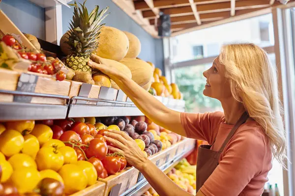 cheerful mature saleswoman smiling and looking happily at fresh pineapple at grocery store