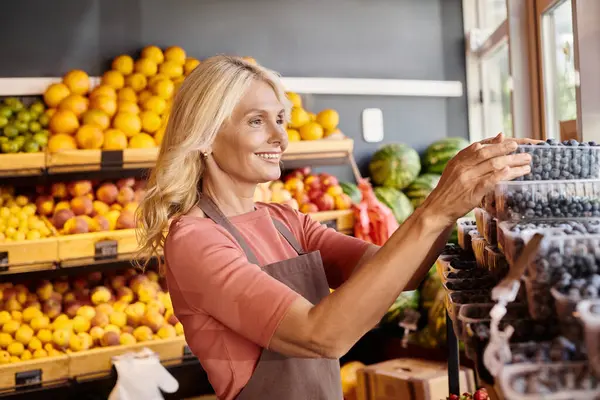 Neşeli olgun satıcı markette arka planda meyve tezgahı olan böğürtlen paketlerini kontrol ediyor.