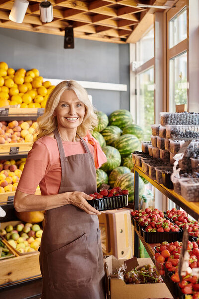 cheerful mature saleswoman holding delicious fresh strawberries and smiling happily at camera