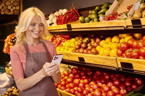cheerful mature saleswoman smiling happily and looking at her mobile phone at grocery store