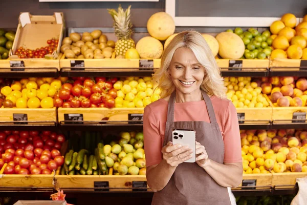 cheerful attractive saleswoman smiling joyfully and looking at her mobile phone at grocery store