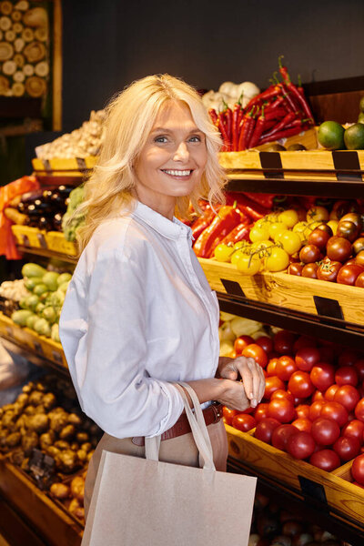 mature jolly customer with paper bag looking happily at camera next to fresh fruits and vegetables