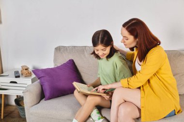 cheerful preteen girl reading special sex education literature near mother at home in living room clipart