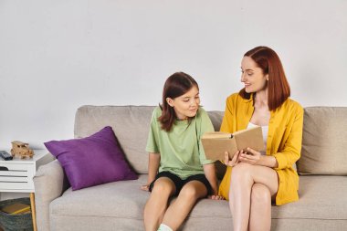 Oturma odasındaki rahat kanepede genç kızına seks eğitimi hakkında kitap okuyan gülümseyen bir kadın.