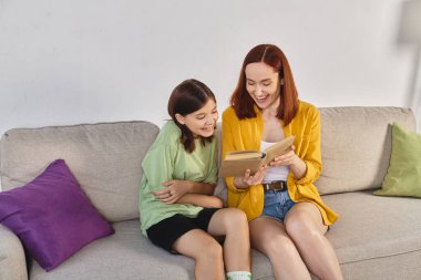 smiling woman reading book about sex education to teenage daughter on cozy couch in living room clipart