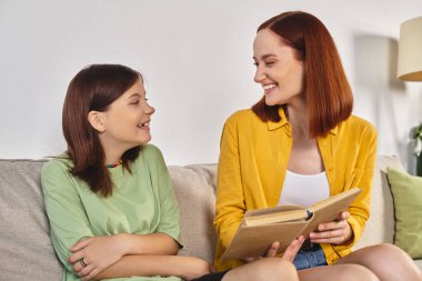 smiling woman reading book about sex education to teenage daughter on cozy couch in living room clipart