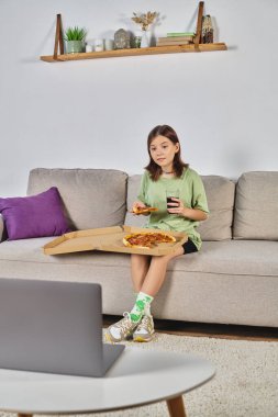 Genç kız elinde pizza ve sodayla kanepede oturuyor ve dizüstü bilgisayarında film izliyor, evde boş vakit geçiriyor.