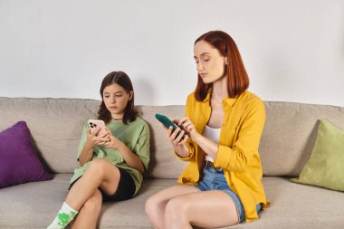 Genç kızı olan yetişkin bir kadın modern oturma odasındaki koltukta otururken akıllı telefon kullanıyor.