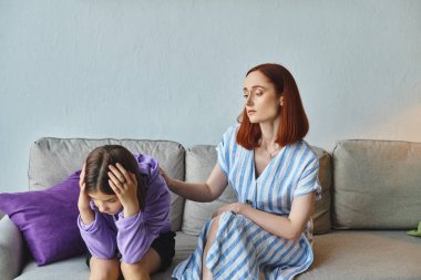 Endişeli bir anne evde başı eğik oturan depresif bir genç kızı sakinleştiriyor.