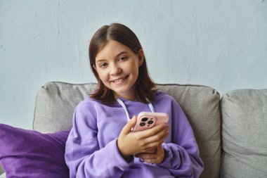 Akıllı telefonuyla kanepede oturan ve oturma odasındaki kameraya bakan neşeli genç kız.