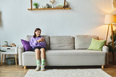 Modern oturma odasındaki rahat koltukta otururken sosyal medyada gezinen neşeli genç kız.