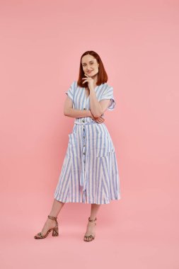 full length of smiling redhead woman in striped dress looking at camera on pink backdrop, fashion clipart