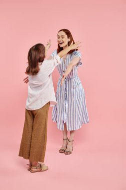 Uzun boylu, neşeli ve şık bir kadın pembe arka planda genç kızına sarılacak.