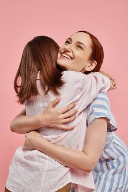 excited and inspired woman embracing teenage girl on pink backdrop, unity of mother and daughter clipart