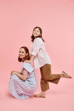 cheerful stylish woman sitting on haunches near teen daughter posing on pink backdrop, fun and joy clipart