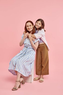 Mutlu genç kız, zarif annesini kucaklıyor. Taş blokta oturuyor. Pembe arka planda, sevgi ve birlik içinde.