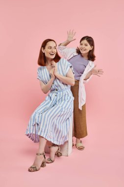 Neşeli genç kız eğleniyor ve annesini korkutuyor. Pembe arka planda taş blokta oturuyor.