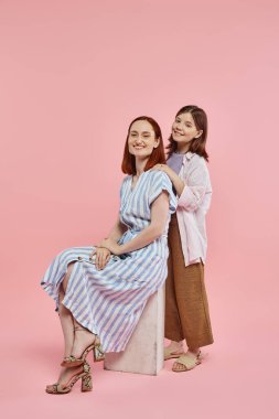 Mutlu genç kız, zarif annesini kucaklıyor. Taş blokta oturuyor. Pembe arka planda, sevgi ve birlik içinde.