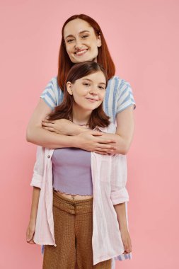 cheerful and stylish woman hugging smiling preteen daughter on pink backdrop, happy family clipart