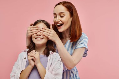 joyful and stylish woman covering eyes of preteen daughter while playing guess who game on pink clipart