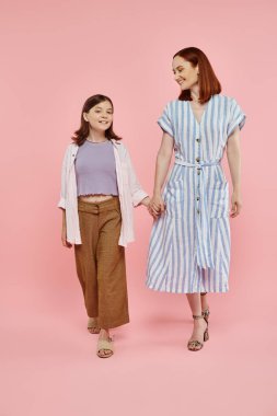 full length of cheerful and stylish woman with preteen daughter holding hands and walking on pink clipart