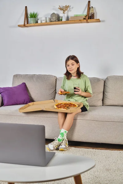teenage girl sitting on couch with pizza and soda and watching movie on laptop, leisure at home