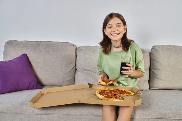 stock image joyful teenage girl looking at camera while sitting on couch with pizza and soda, meal time