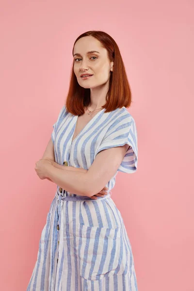 stock image confident woman in striped dress posing with folded arms and looking at camera on pink backdrop