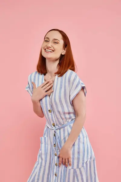 stock image excited redhead woman in trendy striped dress looking at camera and laughing on pink backdrop