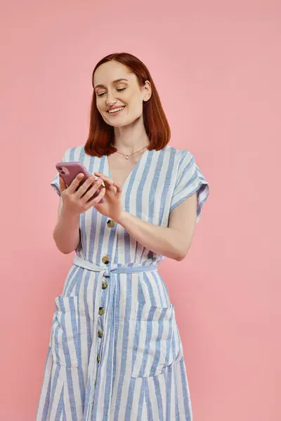 stock image cheerful and stylish woman in striped dress using mobile phone on pink backdrop, modern lifestyle