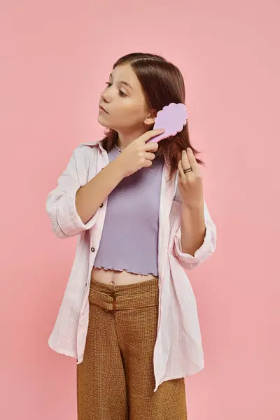 preteen girl in stylish casual attire brushing hair on pink backdrop in studio, beauty routine