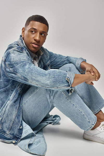 симпатичный молодой африканский американец в модном джинсовом наряде сидит на полу, концепция моды