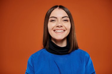 Burnu delinmiş mutlu genç bir kadın kameraya bakıyor ve turuncu arka planda gülümsüyor, mavi ceketli.