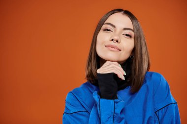 Burnu delinmiş düşünceli bir kadın kameraya bakarken turuncu arka planda, mavi ceketli.