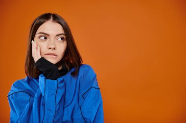 pensive woman with pierced nose biting lip and looking away on orange background, hand near face clipart