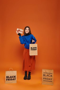 joyful woman holding wrapped present and shopping bag on orange backdrop, black friday sales clipart