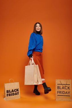 Neşeli bir kadın alışveriş torbaları tutuyor ve neşeli bir şekilde turuncu arka planda yürüyor. Kara Cuma satışları.