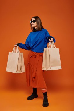 Havalı güneş gözlüklü çekici bir kadın elinde çantalarla turuncu arka planda, siyah cuma satışları yapıyor.