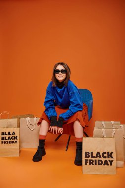Güneş gözlüklü pozitif kadın siyah cuma alışveriş torbalarının yanında koltukta oturuyor.