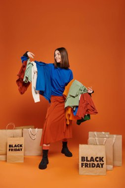 Kumral, mutlu bir kadın. Elinde renkli sonbahar kıyafetleri var. Kara Cuma alışveriş torbalarının yanında.