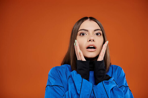 shocked woman with pierced nose touching cheeks with hands and looking at camera on orange backdrop