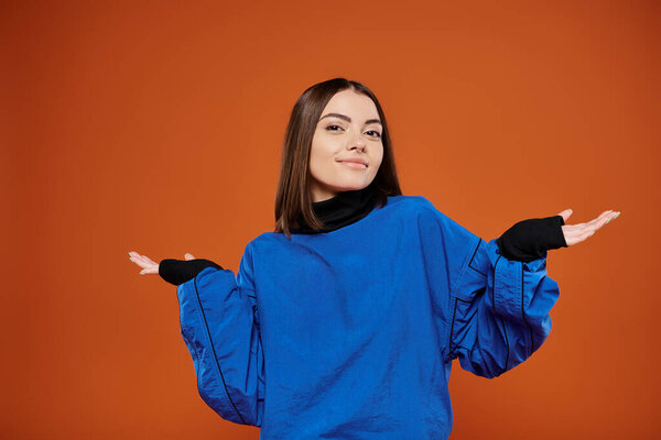 confused woman with pierced nose looking at camera while standing on orange backdrop, blue jacket