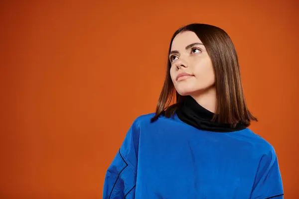 stock image pensive woman with pierced nose looking away while standing in blue jacket on orange background