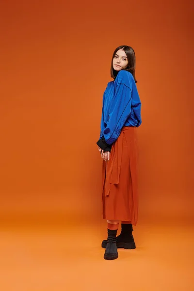 autumn fashion, beautiful woman in skirt, blue sweatshirt and boots standing on orange backdrop