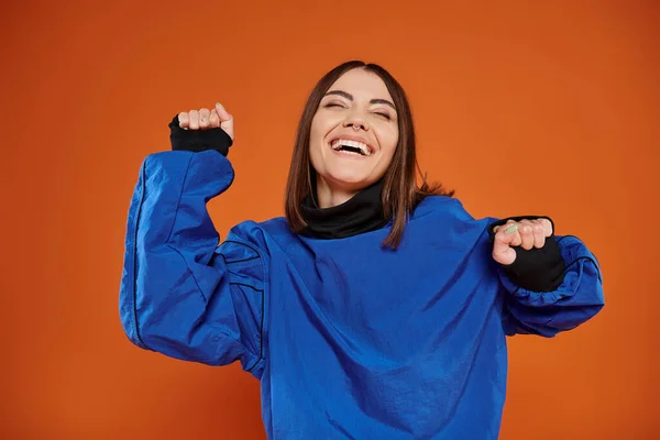 stock image excited young woman with pierced nose gesturing and smiling on orange backdrop, blue sweatshirt