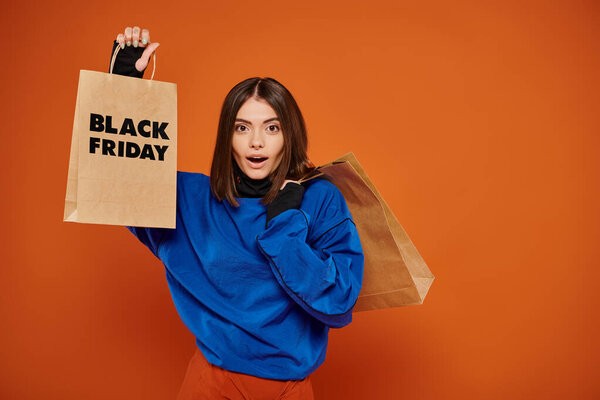 шокированная женщина держит сумки с черными пятничными буквами на оранжевом фоне, сезон продаж
