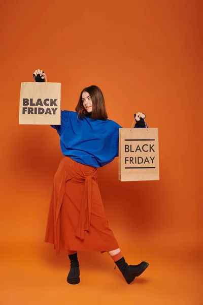 brunette woman in fall attire holding shopping bags with black friday letters on orange backdrop