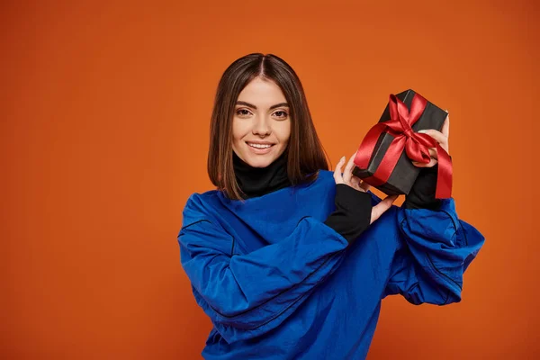 stock image cheerful woman with short brunette hair holding wrapped present on orange backdrop, black friday