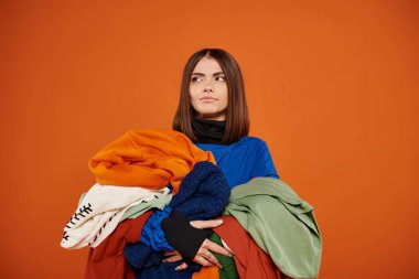 pensive young woman with brunette hair holding pile of colorful autumnal clothes, black friday clipart