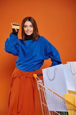 Kredi kartı taşıyan mutlu bir kadın. Turuncu renkli alışveriş çantalarıyla dolu bir arabanın yanında. Kara Cuma konsepti.